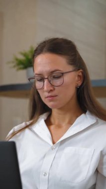 Gözlük takan ve şık giyinen genç bir iş kadını, doğal ışıkla dolu rahat bir oturma odasında çalışırken kahverengi kanepeye oturur ve laptopuna odaklanır. dikey video