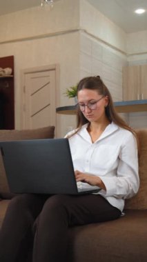 Gözlük takan ve şık giyinen genç bir iş kadını, doğal ışıkla dolu rahat bir oturma odasında çalışırken kahverengi kanepeye oturur ve laptopuna odaklanır. dikey video