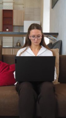 Gözlük takan ve iş kıyafeti giyen genç bir kadın rahat bir şekilde kahverengi koltukta oturur, rahat bir ev ortamında çalışırken bilgisayarında daktilo yazmaya odaklanır. dikey video