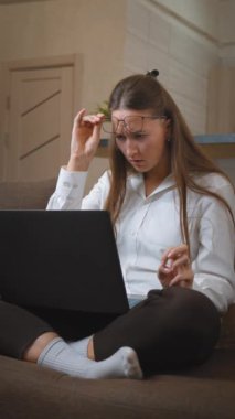 Genç bir kadın dizüstü bilgisayar ekranında kaşlarını çatarken gözlüklerini ayarlıyor. Uzaktaki işlerle uğraşırken kafası karışıyor ve hayal kırıklığı yüzünü yansıtıyor. dikey video