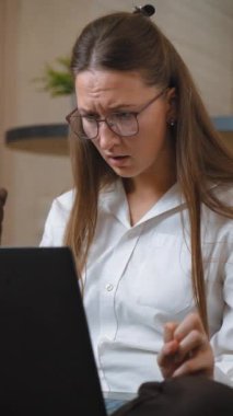 Bilgisayarına uzaktan bakan genç bir iş kadını yüzünde şok olmuş bir ifadeyle ekranda gördüklerine şaşırarak gözlüğünü düzeltiyor. dikey video