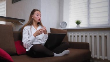 Genç bir kadın koltuğunda dizüstü bilgisayarını kullanarak bacak bacak üstüne atmış oturuyor. El sallıyor, gülümsüyor ve konuşuyor.