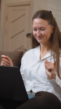 Genç bir kadın evindeki kanepesinde oturuyor, bilgisayarında canlı bir video görüşmesi yapıyor, rahat, modern yaşam alanında sohbet ederken hareketler yapıyor ve gülümsüyor. dikey video