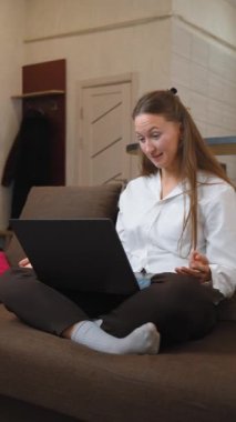 Genç bir kadın evindeki kanepesinde oturuyor, bilgisayarında canlı bir video görüşmesi yapıyor, rahat, modern yaşam alanında sohbet ederken hareketler yapıyor ve gülümsüyor. dikey video