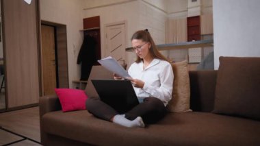 Ofisteki kanepede oturan iş kadını laptopun önündeki belgelere ciddi bir ifadeyle bakıyor. Odaklanmış ve meşgul, evrak işlerini analiz ediyor, son teslim tarihleri ve stresle başa çıkıyor.