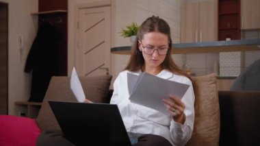 Ofisteki kanepede oturan iş kadını laptopun önündeki belgelere ciddi bir ifadeyle bakıyor. Odaklanmış ve meşgul, evrak işlerini analiz ediyor, son teslim tarihleri ve stresle başa çıkıyor.