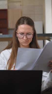 Genç iş kadını kanepede oturuyor, belgeleri gözden geçiriyor ve dizüstü bilgisayarla çalışıyor. Gözlük takıyor ve işine odaklanmış gibi görünüyor. dikey video