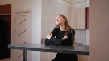 Genç kadın modern mutfakta tezgaha yaslanıyor, siyah elbise giyerken hayal kuruyor. Huzurlu bir tefekkür anının tadını çıkarırken rahatlık boşluğu dolduruyor.