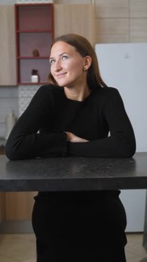 Siyah gömlekli genç bir kadın masada oturuyor, gülümsüyor ve farklı yönlere bakıyor, mutluluk duygusunu somutlaştırıyor ve iç mekanda derin düşüncelere dalıyor. dikey video