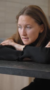 Mutfakta bar taburesinde oturan genç bir kadın başını elinde tutuyor ve baş ağrısıyla boğuşuyor. Acıyla başa çıkarken hayal kırıklığı ve yorgunluk aşikar. dikey video