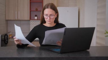 Evinden dokümanlara bakıp dizüstü bilgisayarda yazan genç bir kadın. Odaklanmış ve ciddi bir şekilde mutfak ofisinin rahat atmosferindeki görevleriyle başa çıkıyor.