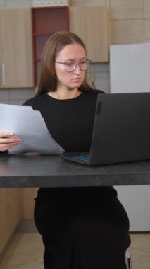 Gözlük takan iş kadını evindeki bilgisayarının başında oturuyor ve ekranındaki verilerle kağıt belgelerindeki bilgileri karşılaştırıyor. dikey video
