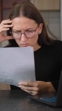 Gözlüklü kadın muhasebeci, evrak okuyor ve masasında dizüstü bilgisayarla çalışırken alnında gözlüklerle geziyor. Belgeleri yönetmede zorluklar yaşıyor.