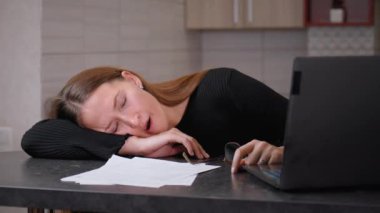 Yorgun bir ofis çalışanı uzun çalışma saatlerinden sonra masasında uyur. Evraklar ve dizüstü bilgisayarlarla çevrili halde gözlerini açık tutmak için çabalarken yorgunluk devreye giriyor.