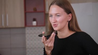 Mutfak masasında oturan genç bir kadın akıllı telefonuyla sesli mesaj kaydederken evde rahat bir anın tadını çıkarırken elsiz bir şekilde konuşuyor.