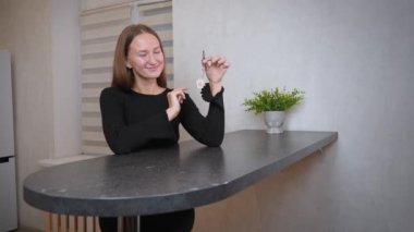 Evinin mutfağında oturmuş gülümseyen genç bir kadın elinde yeni evinin anahtarları, hayatında yeni bir sayfa açmanın heyecanı ve ev sahipliğinin neşesini kucaklıyor.