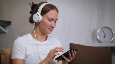 Genç bayan oturma odasında kanepede otururken akıllı telefon kullanıyor. Kulaklık takıyor ve müzik dinliyor.