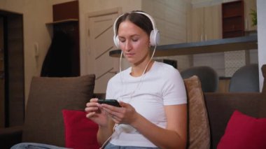 Genç bayan oturma odasında kanepede otururken akıllı telefon kullanıyor. Kulaklık takıyor ve müzik dinliyor.