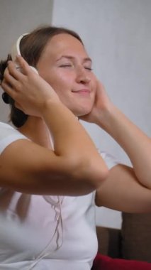 Oturma odasındaki kanepede oturan, kulaklık takan ve müzik dinleyen genç bir kadın. Kafasını ve vücudunu ritme göre hareket ettirir ve elleriyle başını tutar. dikey video