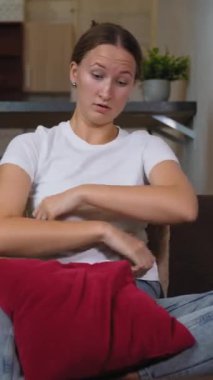 Oturma odasındaki kahverengi kanepede oturan kadın karın ağrısı çekiyor. O, kırmızı yastığı tutuyor ve rahatsızlık içinde ürküyor. dikey video