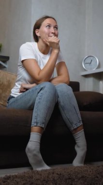 Kanepede oturan genç bir kadın, rahat bir şekilde televizyon izlerken heyecanını ve neşesini dile getiriyor, akşamları oturma odasında, kişisel eğlence ve eğlencenin tadını çıkarıyor. dikey video