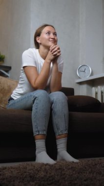 Genç kadın kanepede oturuyor, rahat bir şekilde televizyon izlerken heyecan ve neşe ifade ediyor, akşamları oturma odasında, kişisel eğlence ve eğlencenin tadını çıkarıyor.