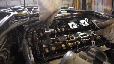 Eldiven giyen tamirci, araba motorunun kapağındaki cıvataları sıkmak için İngiliz anahtarı kullanıyor. Motor açık, şaftlar, vanalar ve zamanlama zincirini gösteriyor.