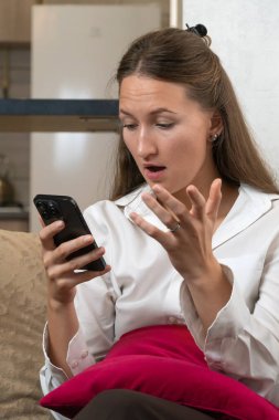 Şaşırmış iş kadını, evdeki bir kanepede rahat bir şekilde oturup şaşkınlık ve inançsızlığını dile getirirken akıllı telefonuyla beklenmedik haberlere tepki veriyor. Dikey fotoğraf