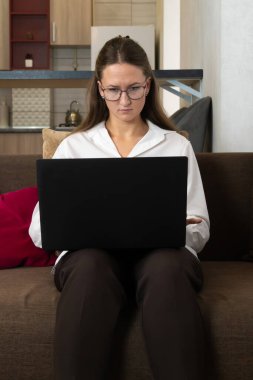 Gözlüklü ve beyaz tişörtlü genç bir kadın şık bir ev ofisteki koltukta rahat bir şekilde oturuyor uzaktan çalışma ve internet iletişimiyle uğraşırken dizüstü bilgisayarına odaklanıyor.
