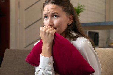 Evdeki kanepede oturan bir kadın, elinde yastıkla tırnaklarını yiyor. Stres ve bunaltıcı duygularla boğuşurken endişe ve korkuyu açığa vuruyor.