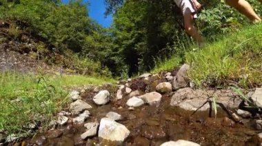 İki yürüyüşçü, açık mavi gökyüzünün altında canlı bir ormanda kayalık dereyi aşarak açık hava maceraları ve doğal keşifler sergiliyorlar.