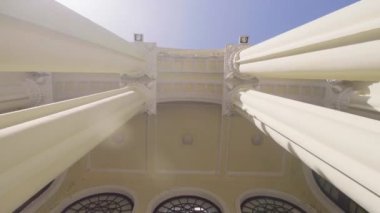 Mimari zarafet, uzun sütunlarla süslü bina cephesini gözler önüne seriyor. Kemerli girişler. Ve dekoratif tavanları vurgulayan iç detaylar. Kamera dikey olarak yukarı yönelir