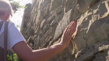 Güneş ışığı, eski taş duvarların dokularına nazikçe dokunurken ve onları incelerken kadının elini aydınlatıyor ve tarih ve doğa ile bir bağ kuruyor. Elinle eski duvara dokunuyorsun..