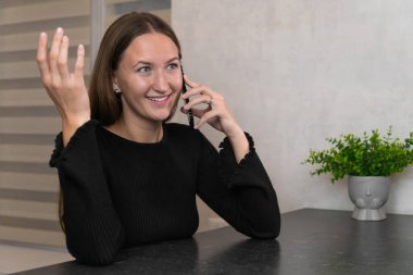 Mutlu genç kadın bir masada oturuyor, telefonuyla konuşuyor ve elleriyle el kol hareketi yapıyor, evinin rahatlığında keyifli bir sohbetin tadını çıkarıyor.