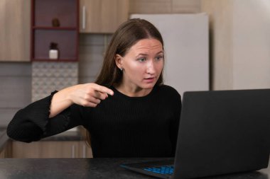 Mutfak tezgahında oturan genç bir kadın, dizüstü bilgisayarı işaret ederken şaşkın şaşkın bakıyor. Uzak çalışma deneyimini yansıtan şaşkınlık ve endişe duyguları.