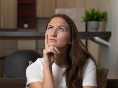 Genç bir kadın evdeki bir kanepede oturuyor, eliyle çenesine dokunuyor ve dikkatlice yukarı bakıyor, bir karar ya da sorun üzerinde düşünüyor.