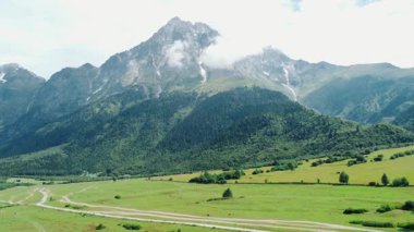 Beyaz bulutlar nazikçe yükselen dağ zirvesini okşar, aşağıdaki parlak yeşil vadiye gölgeler bırakır, toprak yol manzaranın içinden eser. İHA 'dan ateş ediliyor.