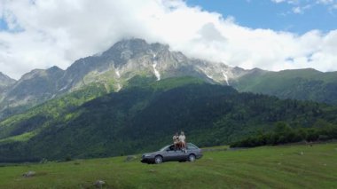 MESTIA, GEORGIA - 2 Temmuz 2024: Neşeli gezgin erkek ve kadın arabanın çatısında oturmuş manzaralı dağ manzarasının keyfini çıkarıyorlar özgürlüğün ve yolculuk paylaşımının vücut bulmuş hali. İHA 'dan ateş.