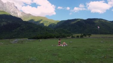 Güneşli bir günde güneşli bir dağ vadisinde, yemyeşil otların arasında, serin bir battaniyenin üzerinde oturan iki kadın turist, çarpıcı yüksek tepelerin arka planında İHA görüntüleriyle eğleniyor..