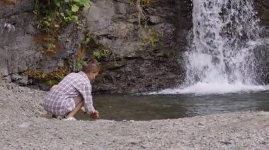 Pembe ekose gömlekli bir kadın ormanda şelalenin yanındaki kayalık derede ellerini yıkıyor. Doğayla olan bağını ve sakinliğini gösteriyor.