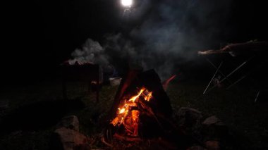 Kamp ateşinin odunları ve parlayan közleriyle yakın plan, parlak alevler karanlık arka planda yanıp sönüyor, sıcak ve rahat bir atmosfer yaratıyor.