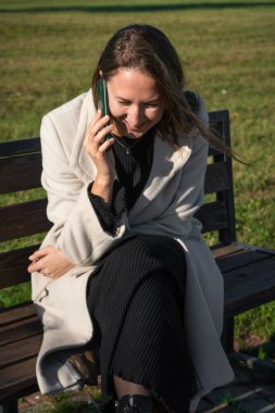 Genç, gülümseyen bir kadın, parktaki bankta oturup güneşli bir sonbahar gününün tadını çıkarırken akıllı telefonuyla mutlu bir şekilde konuşuyor. Dikey fotoğraf