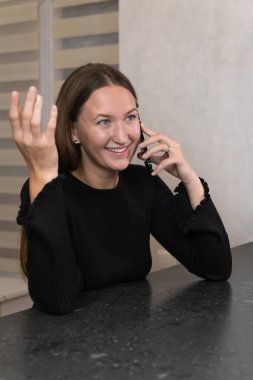 Cep telefonuyla konuşan mutlu genç bir kadın modern dairesinde mutfak tezgahında oturmuş keyifli bir sohbetin tadını çıkarıyor. Dikey fotoğraf