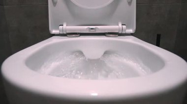 Modern banyodaki temiz su akışı ve beyaz seramik tuvalet sifonunun koyu gri fayanslarla kaplı olması banyonun temizlik ve sterilizasyon sürecini gösteriyor. Yavaş çekim.