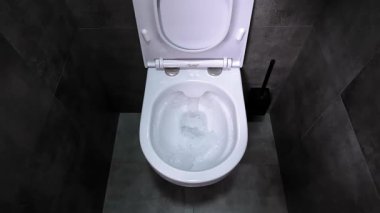 Pürüzsüz, beyaz seramik klozetin içinden akan güçlü su akıntısı. Etrafı koyu gri fayanslı banyo döşemeleri ile çevrili. Modern ev temizliğini gösteriyor. yavaş çekim