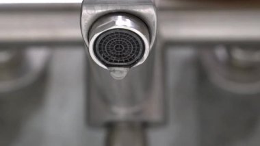 Musluğun koyu gri metal oluğundan su damlacıkları oluşur ve düşer, iç ortamlardaki çevresel su atıkları ve kaynak koruma sorunlarını vurgular