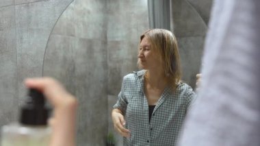 Genç bir kadın saç ürünü uyguluyor. Yuvarlak aynanın önünde dikkatlice ıslak saçlarını şekillendiriyor. Özel bakım sırasında modern banyo ortamında.