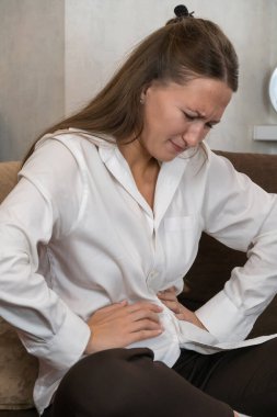 Evdeki kanepesinde oturan genç bir kadın, muhtemelen regl, apandisit veya gıda zehirlenmesi nedeniyle şiddetli karın ağrısı çekiyor. Dikey fotoğraf