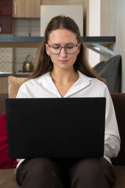 Gözlüklü ve beyaz gömlekli genç bir iş kadını evdeki bir kanepede oturuyor, bilgisayarının üzerinde çalışmaya odaklanmış, uzaktan çalışmanın esnekliğinin tadını çıkarıyor. Dikey fotoğraf