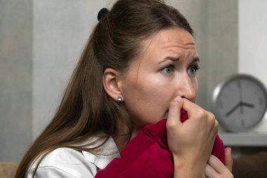 Stresli genç bir kadın kırmızı yastığı tutuyor ve gergin bir atmosferle çevrili parmaklarını endişeyle ısırıyor, işyerinde endişe ve duygusal gerginliği somutlaştırıyor.